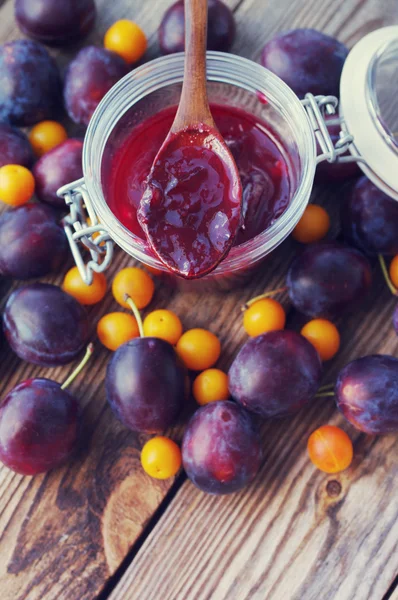 Plum jam in a glass jar and a wooden spoon, and plums with a yellow cherry plum on a wooden surface.