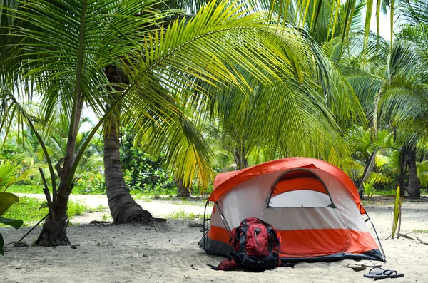 White-orange summer tent, red backpack and slippers in coconut palm tree forest next to sandy beach