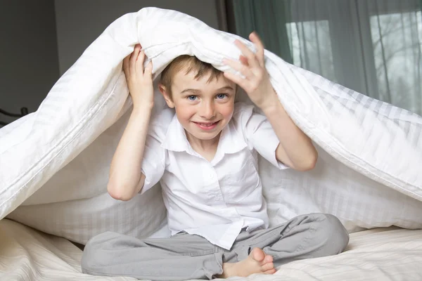 Smiling boy hiding in bed under a white blanket or coverlet.