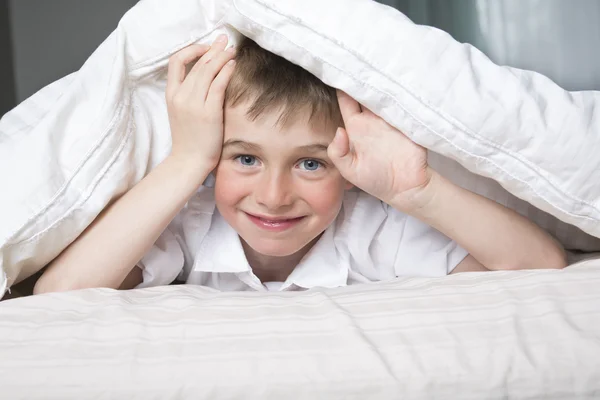 Smiling boy hiding in bed under a white blanket or coverlet.