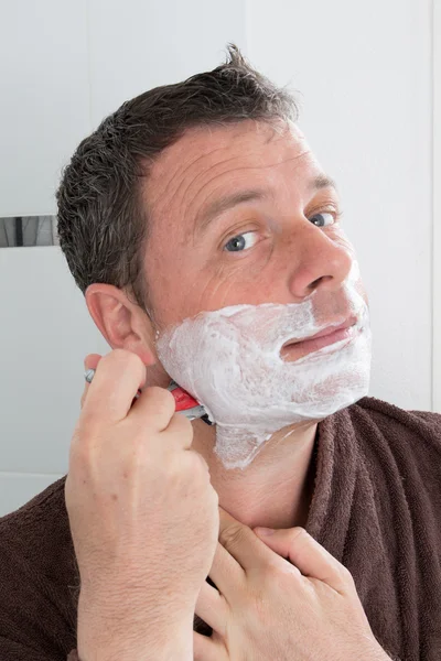 Man shaving in the bath. He is passing the razor for the beard while it looks at the mirror