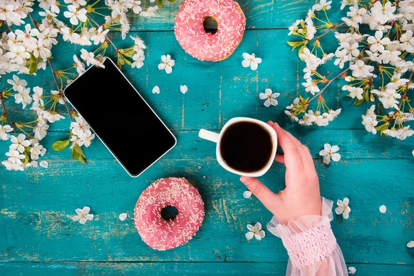 The concept with morning coffee in a romantic style on the wooden background. Cherry blossoms, donuts, smartphone, coffee and hand in the frame.