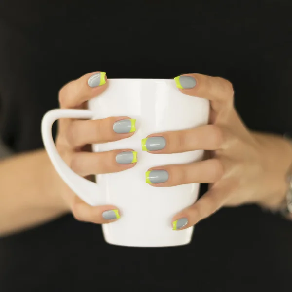 Woman in the black sweater holding white cup in her hands with a bright gray and green nail polish made french style, closeup.