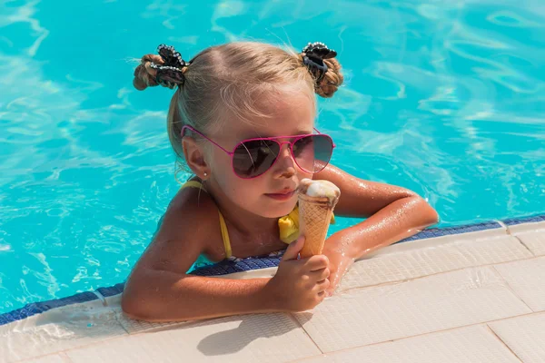 On a sunny day, a cheerful little girl in sunglasses sitting in the pool and eating ice cream