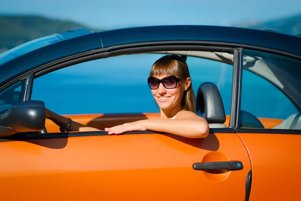 Beautiful young woman with long hair sitting and smiling in orange cabriolet at the Mediterranean sea coast