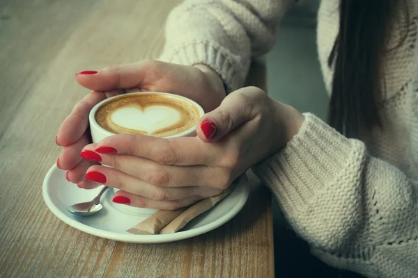 Close-up female hands holding cup with coffee cappuccino with foam with pattern heart. Perfect red gel polish manicure. Wood natural table. Creative color warm post processing instagram style.