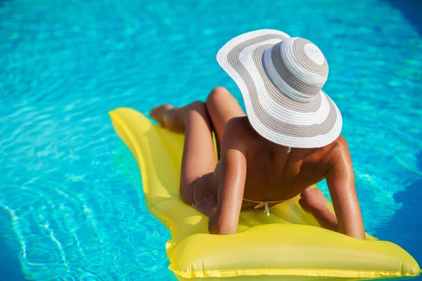 Portrait of beautiful tanned woman relaxing in swimming pool in white swimwear, hat and sunglasses. Exotic model. Creative gel polish manicure and pedicure. Hot summer day and bright sunny light.