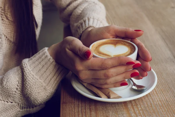 Close-up female hands holding cup with coffee cappuccino with foam with pattern heart. Perfect red gel polish manicure. Wood natural table. Creative color warm post processing instagram style.