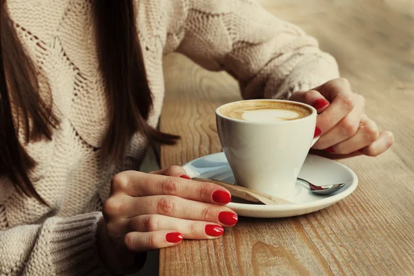 Close-up female hands holding cup with coffee cappuccino with foam with pattern heart. Perfect red gel polish manicure. Wood natural table. Creative color warm post processing instagram style.