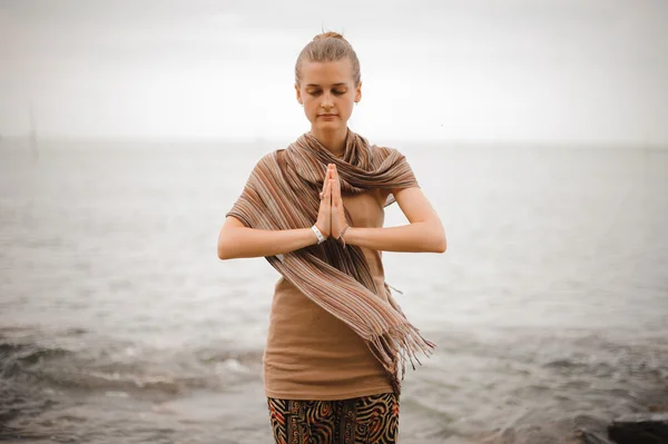 Woman standing a grateful namaste yoga pose on the beach next to the ocean in cloudy weather. Zen, meditation, peace.