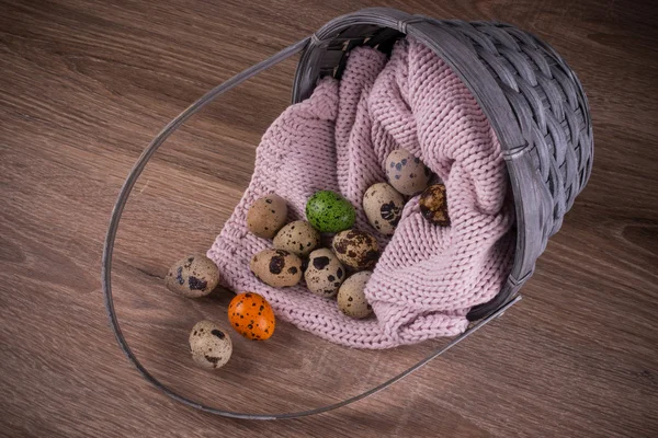 Quail eggs and one orange , green egg spilling out of basket with pink textile on wooden  background
