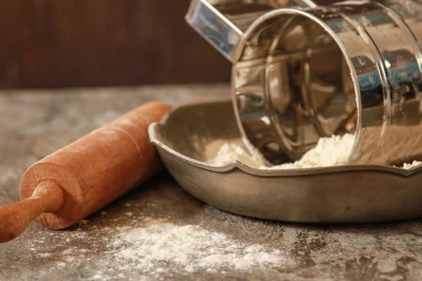 Flour in a metal bowl with  wooden rolling pin on  stone table.
