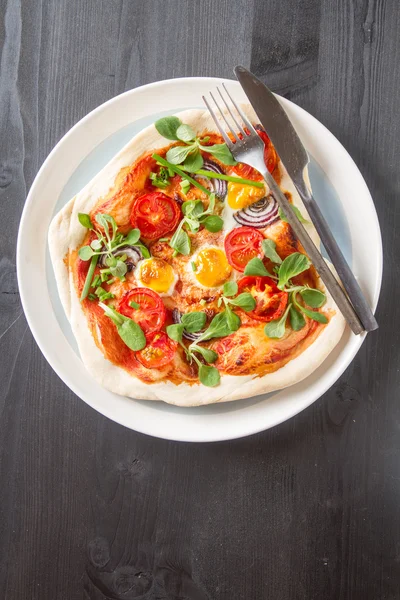 Home Italian pizza with tomato and quail eggs salad on a white plate. Black wooden table. Top view