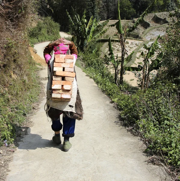Old woman heavily loaded with stack of bricks on her back walks along rural path