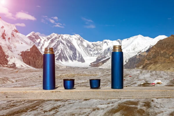 Two Blue Thermoses and Cups on Wooden Table with Mountains on Background and Shining Sun