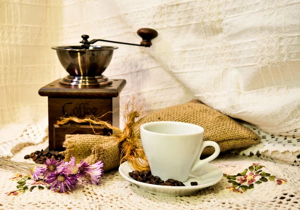 Coffee mill with burlap sack of roasted beans and white cup of prepared espresso on the white knitted linen table-cloth