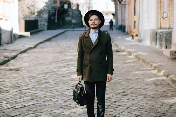 Handsome young bearded hipster man guy in hat Fedora on street with suitcase. Retro vintage fashion look