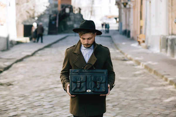 Handsome young bearded hipster man guy in hat Fedora on street with suitcase. Retro vintage fashion look