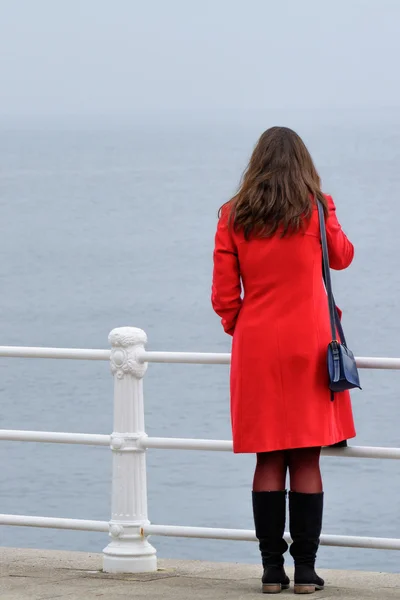 Woman in red coat with black boots sitting in front of seashore.