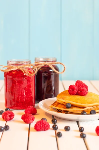 Stack of wheat golden pancakes or pancake cake with freshly picked raspberries on a dessert plate, glass mason jar full off blueberry and raspberry jam.