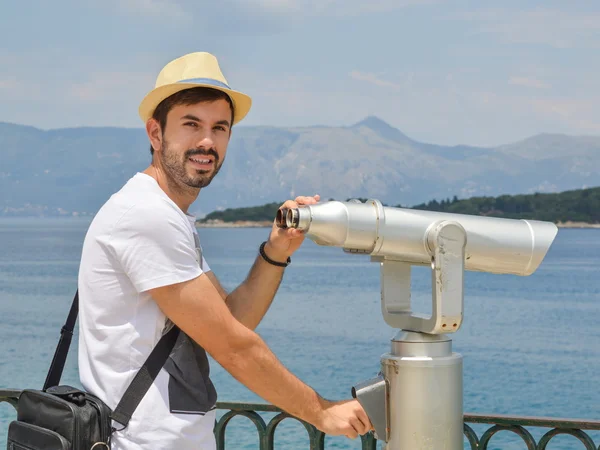Young man holding public binoculars at the seaside wearing straw