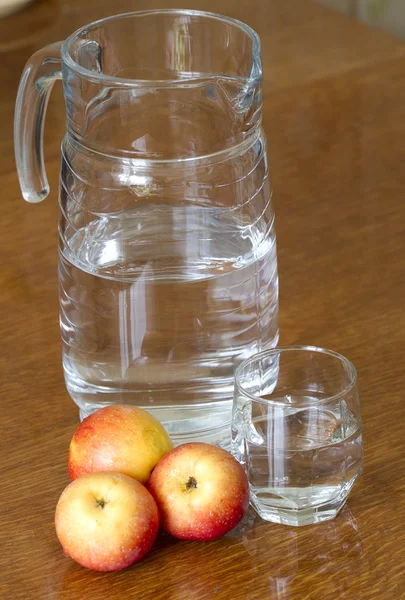 Glass carafe and a glass of water on a wooden table with apples.