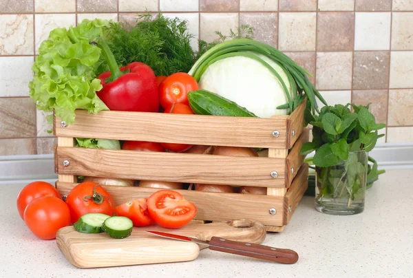 The fresh washed-up vegetables in a wooden box and the cut vegetables on a chopping board against modern kitchen.