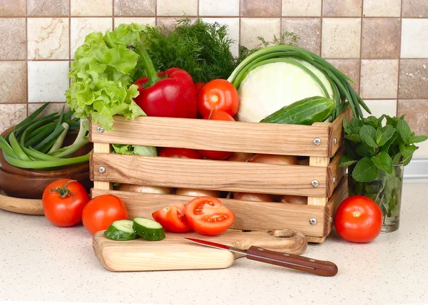 The fresh washed-up vegetables in a wooden box and the cut vegetables on a chopping board against modern kitchen.