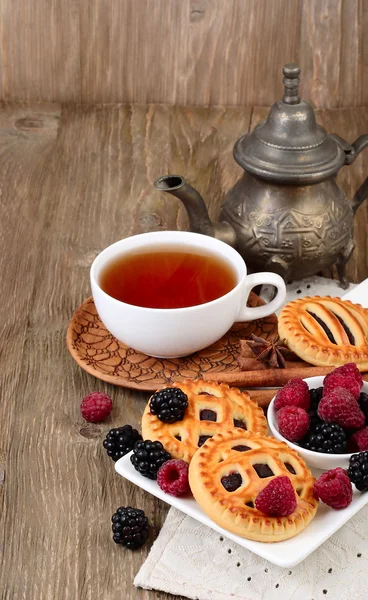 Cup of black tea, berry cookies and fresh raspberry and blackberry on a dark wooden background. Vertical format.