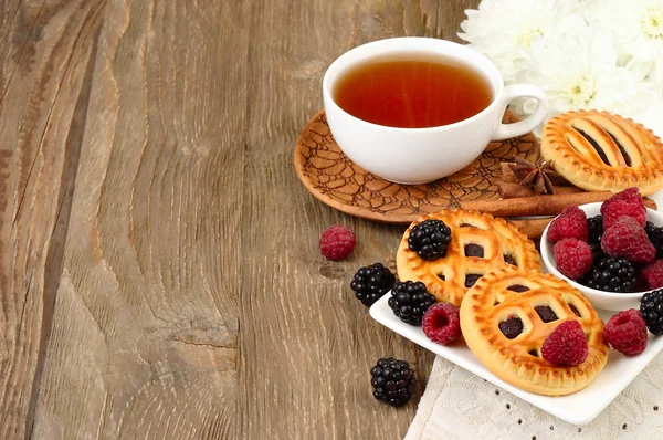 Cup of black tea, berry cookies and fresh raspberry and blackberry on a dark wooden background. Horizontal format.