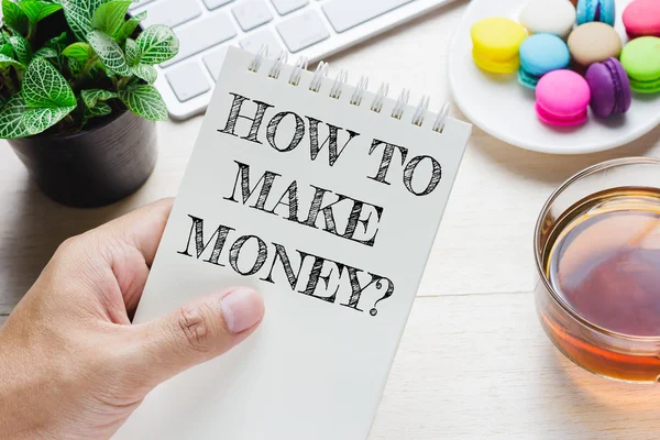 Man holding HOW TO MAKE MONEY message on book and keyboard with a hot cup of tea, macaroon on the table. Can be attributed to your ad.