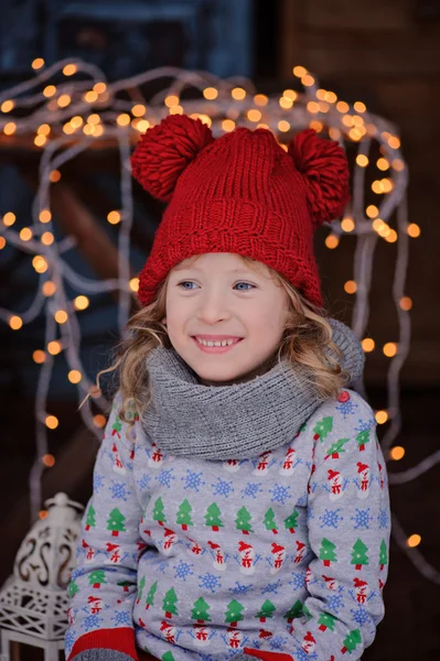 Cute child girl in christmas sweater and red hat sitting outdoor with lights and wooden candle holder