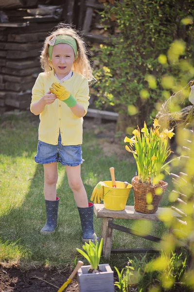 Cute child girl in yellow cardigan plays little gardener and wearing garden gloves in spring garden