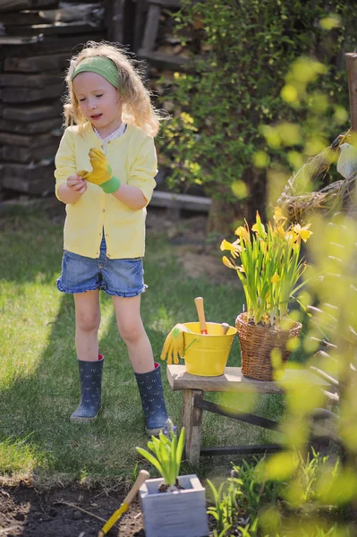 Cute child girl in yellow cardigan plays little gardener and wearing garden gloves in spring garden