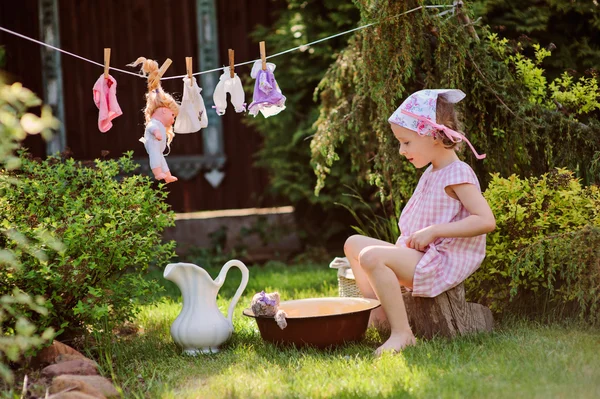 Cute happy child girl in pink plaid dress and flower headband playing toy wash in summer sunny garden