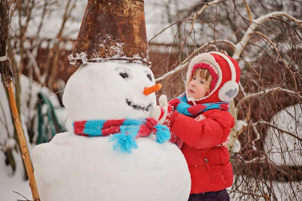 Child girl in red coat making snowman in winter garden