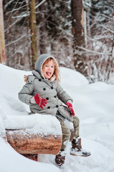 Cute funny child girl in winter snowy forest sitting on wooden log and showing tongue