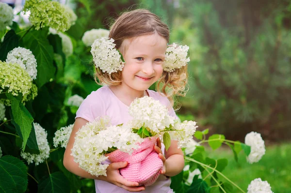 Cute happy child girl playing with hydrangea flowers in summer garden near flowering bush