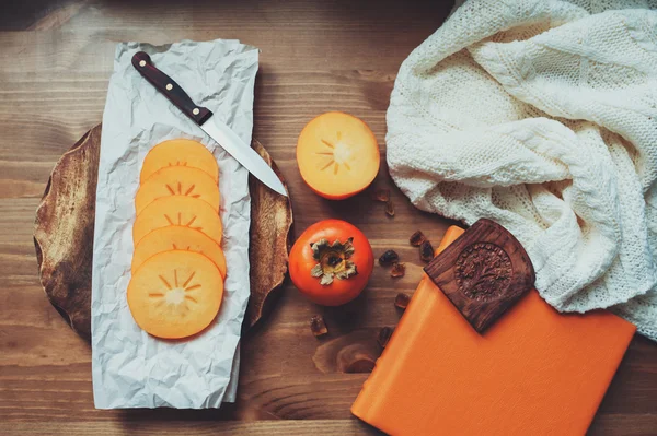 Fresh persimmon slices on wooden table with notebook and knitted sweater, top view, cozy lifestyle setting