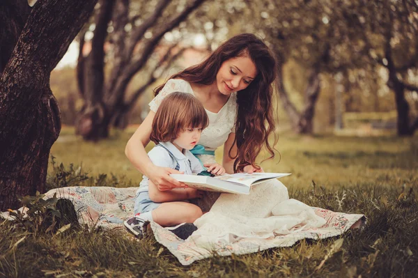 Mother reading book to toddler son outdoor in spring garden. Family spending time together on picnic. Parent teaching kids on summer vacation.