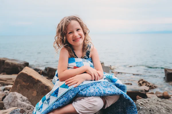 Cute curly happy child girl relaxing on stone beach, wrapped in cozy quilt blanket. Summer vacation activities, traveling in Europe on holidays.
