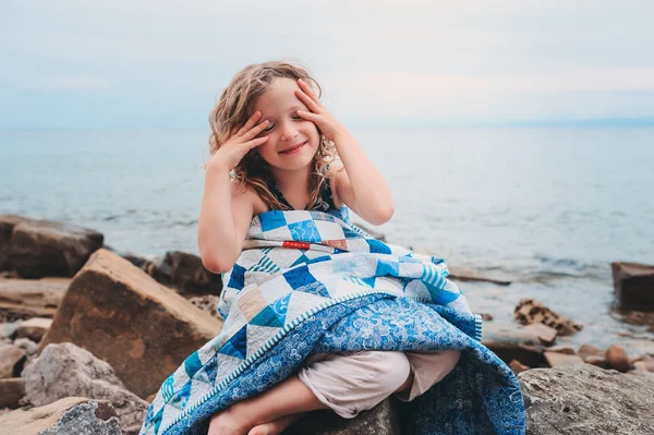 Cute curly happy child girl relaxing on stone beach, wrapped in cozy quilt blanket. Summer vacation activities, traveling in Europe on holidays.