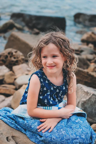 Cute curly happy child girl relaxing on stone beach, wrapped in cozy quilt blanket. Summer vacation activities, traveling in Europe on holidays.