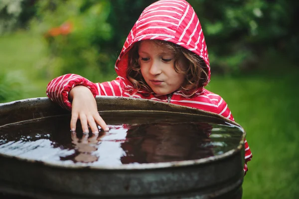 Child girl in red raincoat playing with water barrel in rainy summer garden. Water economy and nature care concept