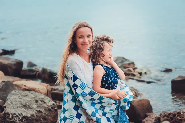 Happy mother and daughter wrapped in quilt blanket spending time together on the beach on summer vacation. Happy family traveling, cozy mood.
