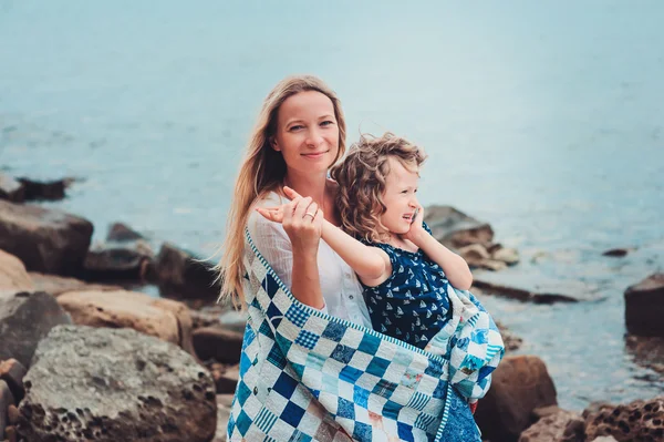 Happy mother and daughter wrapped in quilt blanket spending time together on the beach on summer vacation. Happy family traveling, cozy mood.