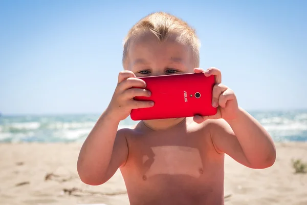 Cute boy laughing while playing with smart phone at beach near the sea