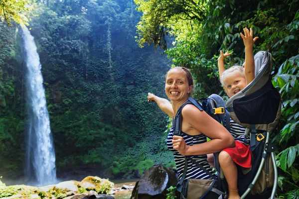 In waterfall pool young happy mother hold little traveller on back - baby girl in carrying backpack enjoying travel adventure, Hiking activity with child on family summer vacation, weekend nature tour