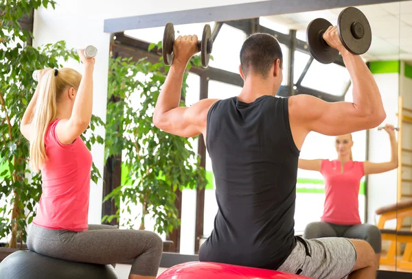 Young couple training in gym with dumbbells in front of a mirror sitting on fitballs