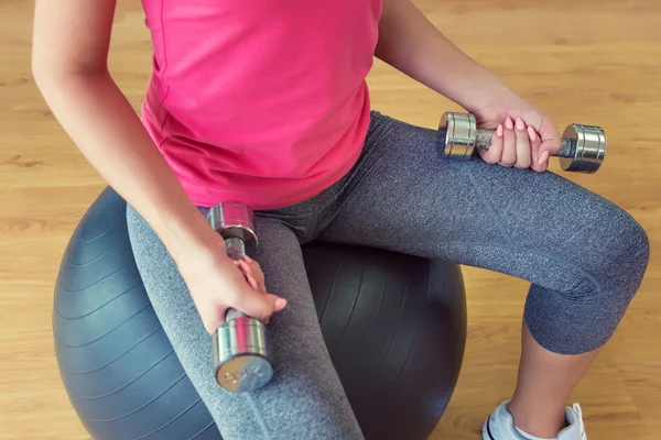 Young woman training in gym with dumbbells in front of a mirror sitting on fitball